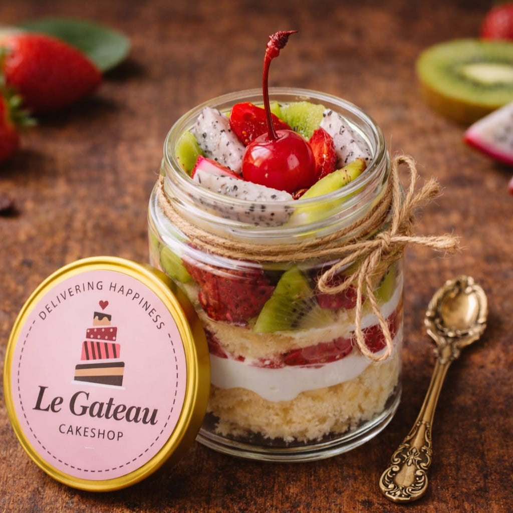 Layered dessert in a jar with fruit and a cherry, branded 'Le Gateau Cakeshop', on a rustic background.
